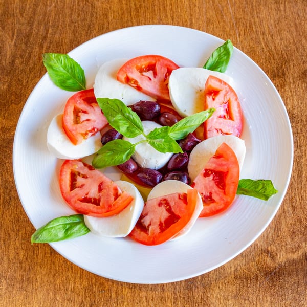 San Vito's Caprese salad with sliced tomatoes, mozzarella, olives, and basil on a white plate.