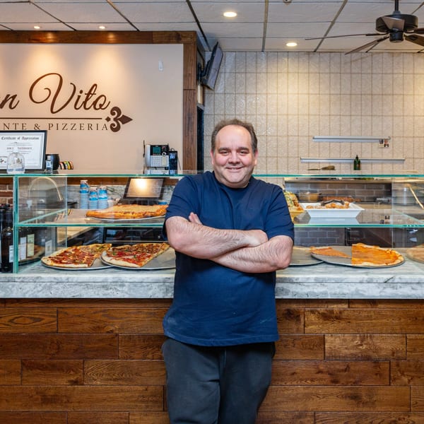 Smiling man with crossed arms stands in front of a counter with San Vito's pizzas.