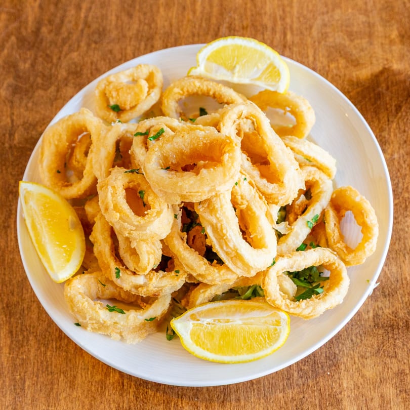 A white plate filled with golden-brown fried calamari rings, garnished with fresh parsley and lemon wedges.